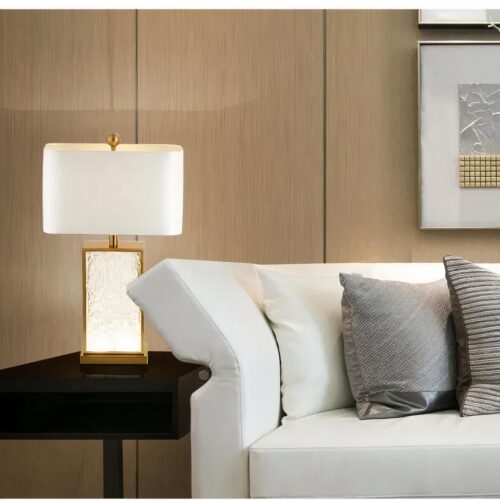 Photo d'une lampe décorative au pied en verre et abat-jour en tissu blanc posée sur un guéridon en bois près d'un canapé blanc moderne