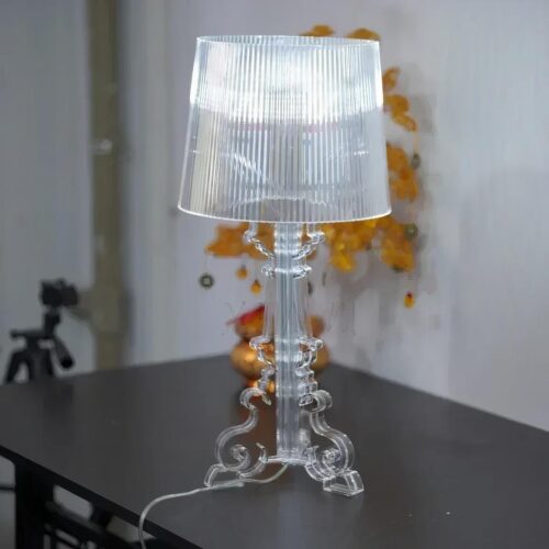 Photo d'une lampe à poser originale transparente sur une table noire devant un bouquet de fleurs jaunes et un mur gris