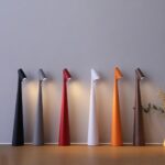 Photo de six lampes sans fil de couleur différentes allumées et posées sur un meuble marron clair devant un mur gris