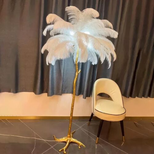 Photo d'une lampe de salon à plumes blanches et pied doré comme une patte d'oiseau posée sur un sol en marbre marron foncé devant un fauteuil blanc et des rideaux noirs