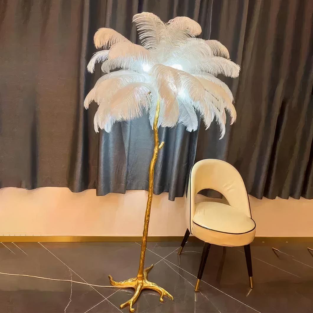 Photo d'une lampe de salon à plumes blanches et pied doré comme une patte d'oiseau posée sur un sol en marbre marron foncé devant un fauteuil blanc et des rideaux noirs
