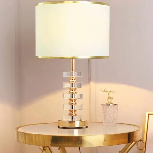 Photo d'une lampe design allumée à l'abat-jour en tissu blanc et au corps doré et cristal posée sur une petite table ronde style guéridon, dorée également, à côté d'une décoration en cristal au couvercle doré en forme de cerf dans un décor beige