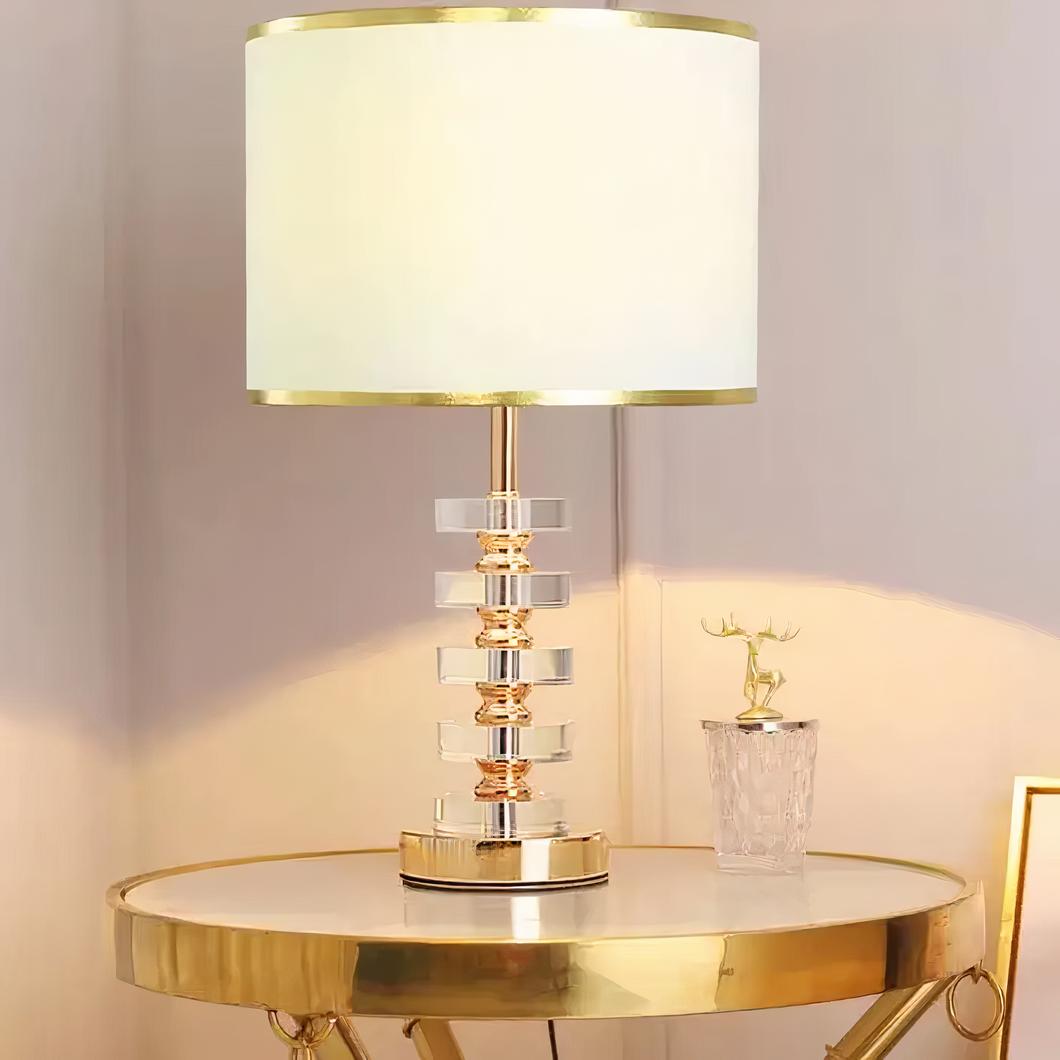 Photo d'une lampe design allumée à l'abat-jour en tissu blanc et au corps doré et cristal posée sur une petite table ronde style guéridon, dorée également, à côté d'une décoration en cristal au couvercle doré en forme de cerf dans un décor beige