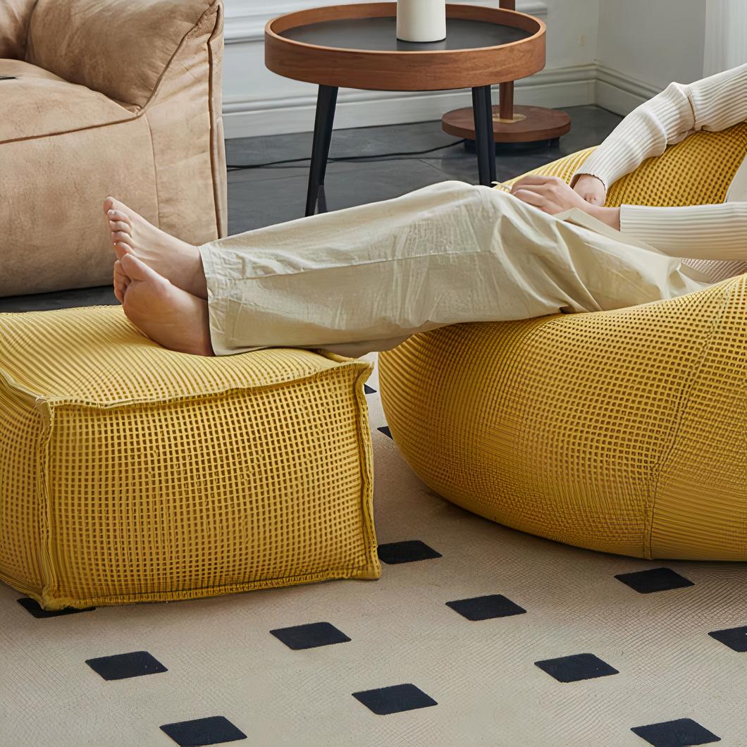 Photo d'un pouf repose pied jaune en tissu soutenant la jambe d'une femme assise sur un canapé assorti, sur un tapis beige et noir